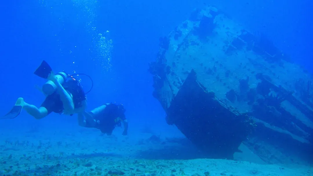 Two scuba divers exploring a sunken shipwreck resting on the sandy seabed