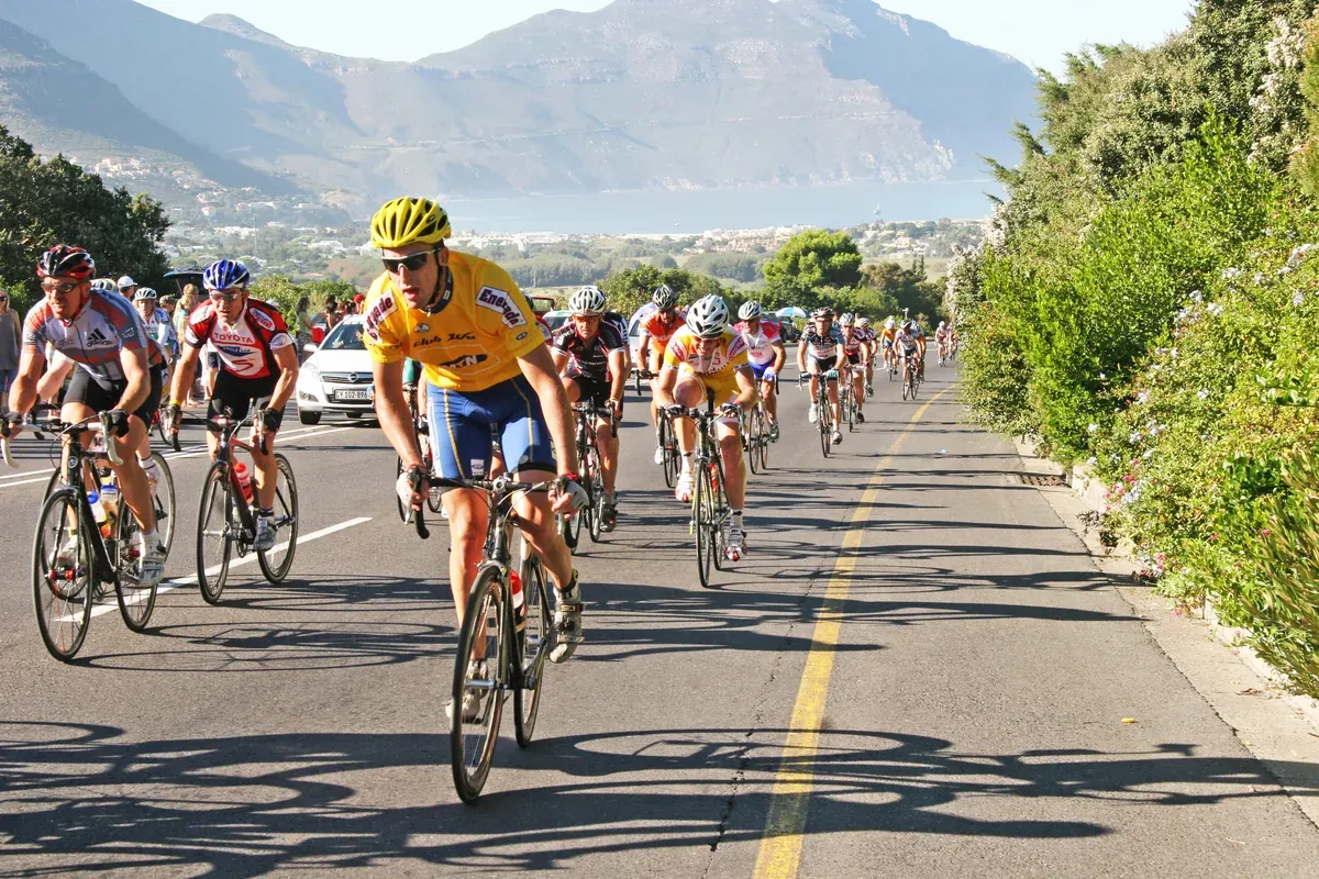Amateur cyclists riding in a peloton on the Cape Town Cycle Tour route, with the Cape Peninsula mountains and ocean in the background.