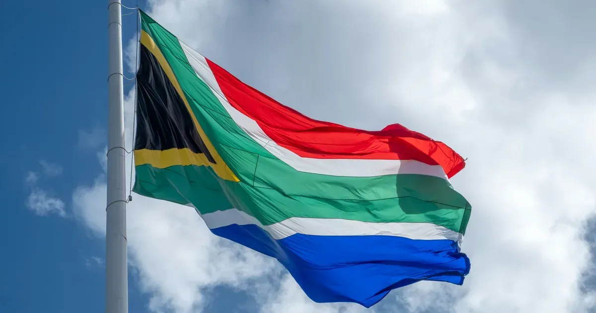 South African flag flying against a blue sky with clouds, illustrating an article on South Africa's narrow tax base and social grant redistribution.