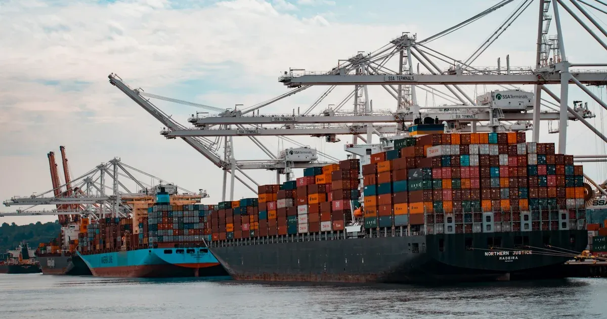 Stacked shipping containers loaded onto container ships at a busy port, gantry cranes overhead